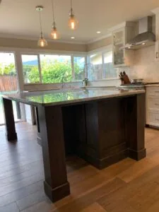 Modern kitchen with dark wood island, glass countertop, pendant lights, and views through large windows to landscaped outdoor space