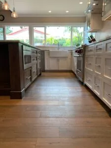 Modern kitchen with white cabinetry, stainless steel appliances, farmhouse sink, pendant lighting, and wood flooring overlooking outdoor views through large windows.