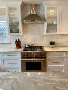 Modern kitchen with white cabinetry, marble countertops, stainless steel range hood, and gas cooktop with red knobs