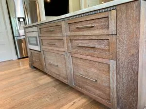 Kitchen island with weathered wood cabinetry, multiple drawers with metal handles, and stainless steel appliances visible in background