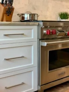 Modern kitchen counter with white cabinetry on left, stainless steel oven with red knobs on right, knife block and potted plant on countertop