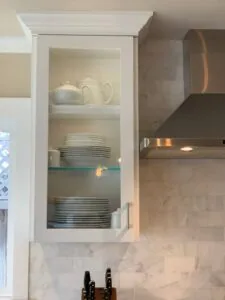 White kitchen cabinet with glass door displaying stacked plates, teapot, and pitcher on shelves