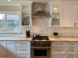 Modern kitchen with white cabinetry, marble countertops, black gas range with red knobs, marble subway tile backsplash, and bronze range hood
