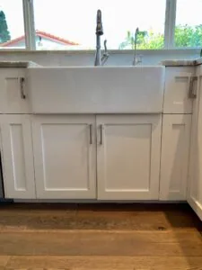 White farmhouse sink with black faucet mounted above white cabinet drawers in kitchen with wood flooring and large window