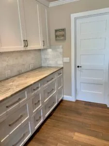White kitchen cabinetry with marble countertops and backsplash, white door, and wood flooring