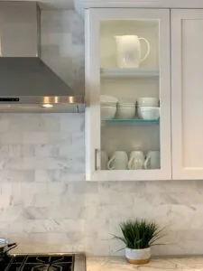 Kitchen cabinet with glass doors displaying white ceramic dishware and pitcher, marble subway tile backsplash, stainless steel range hood, potted plant on counter