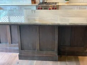 Kitchen island with light marble countertop and dark gray upholstered panel seating below, white cabinetry and gas range visible in background