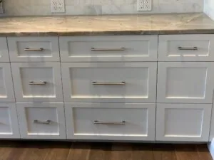 White kitchen cabinetry with brushed metal handles and light marble countertop, white subway tile backsplash with electrical outlets above