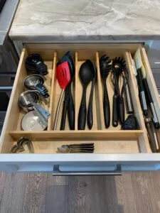 Kitchen drawer organizer with wooden dividers containing cooking utensils, serving spoons, a red spatula, black measuring cups, and kitchen tools neatly arranged in sections